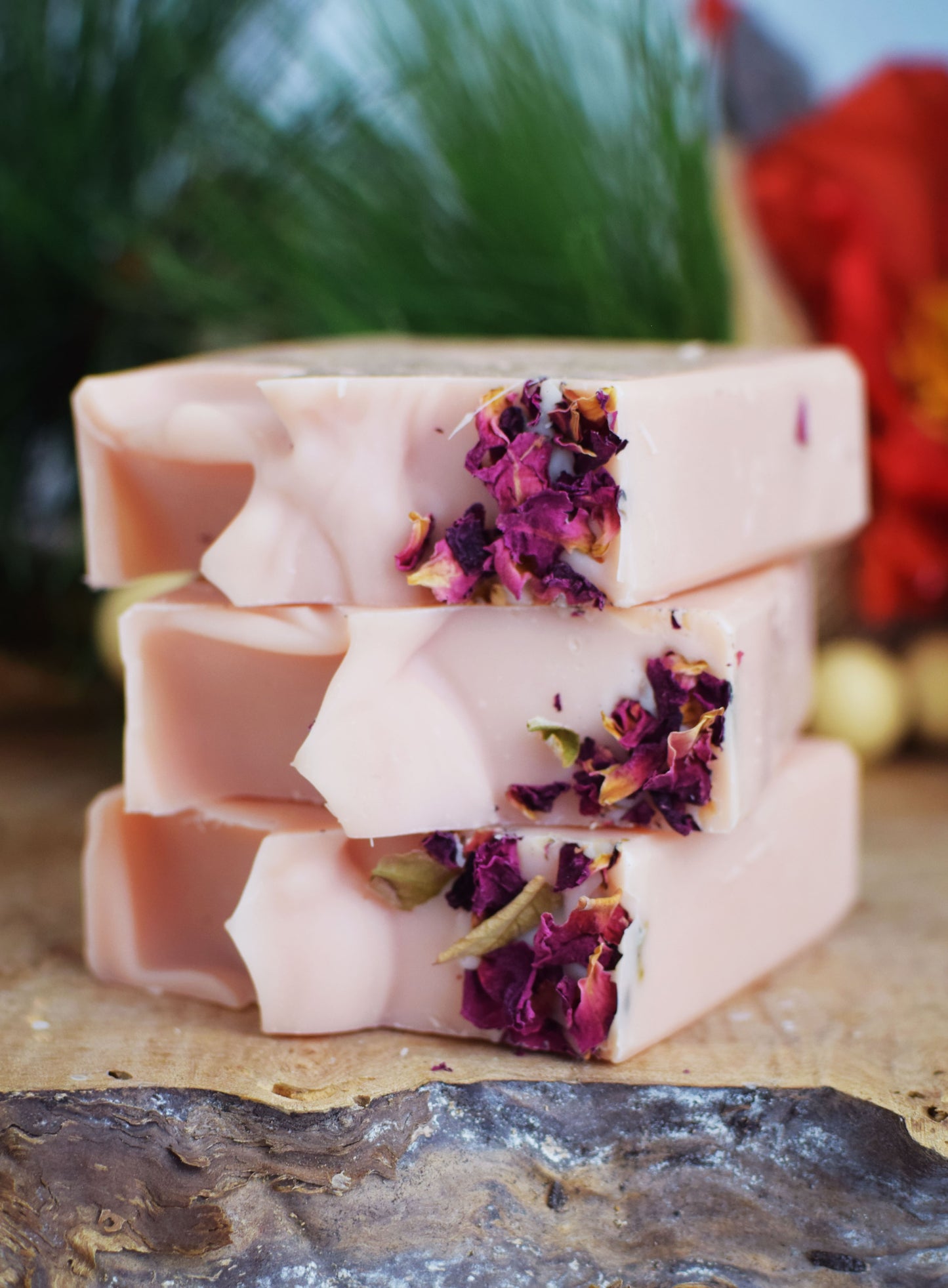 Stack of pink bar soaps with dried rose petals embedded within the soap, placed on a natural looking surface with foliage in the background.