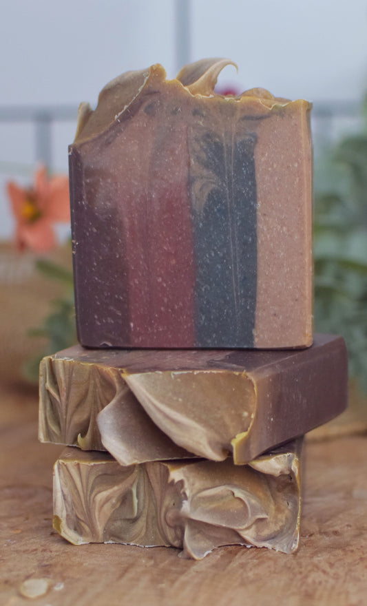 dark brown red dark blue and tan striped soap bar stacked in a pile on a wood surface with a green background