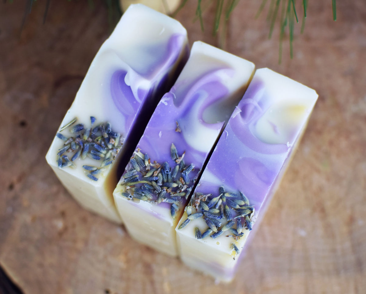 three bars of lavender soap, purple and white swirls with lavender but topping. sitting on a wood surface with pine needles in the background