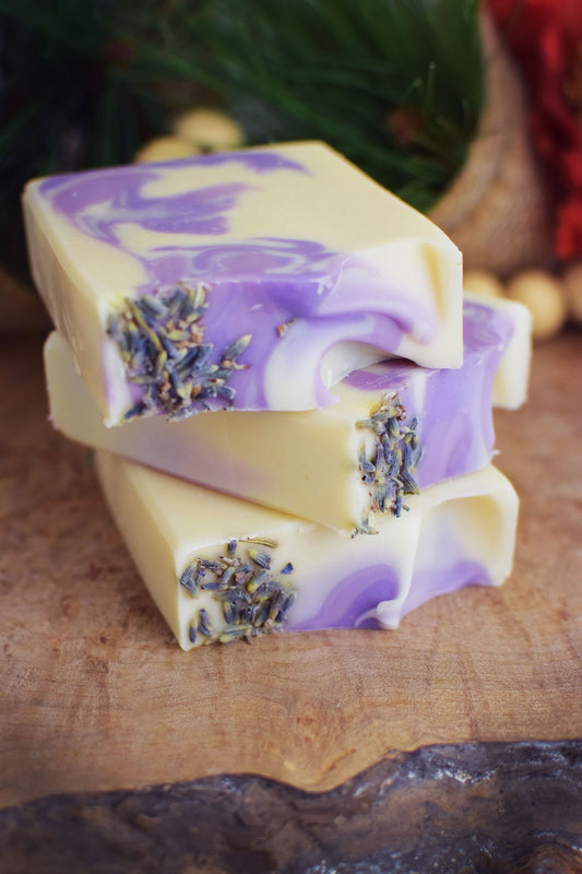 3 bars of lavender soap with a white and purple marbled design, adorned with lavender petals, on a wooden surface with evergreen branches in the background. 